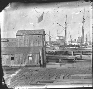 Two story wood framed boathouse in foreground with docks and ships in background at the Detroit River. Recorded in glass negative ledger: "LD/Water supply."